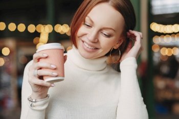 Cropped shot of pleasant looking woman poses in outdoor cafe, holds paper cup of coffee, dressed in white outfit, smiles happily, has talk with colleague or friend during break. Female with beverage