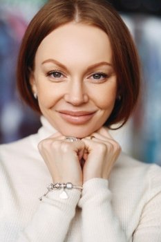 Vertical shot of pleasant looking lady with brown hair, keeps hands under chin, has healthy skin, make up, wears bracelet and white jumper, looks at camera, has broad forehead, nice appearance