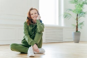 Healthy lifestyle concept. Beautiful slim young woman with red wavy hair, keeps hand under chin, smiles happily, dressed in sportswear, has flexible body, sits on floor. Female has home workout