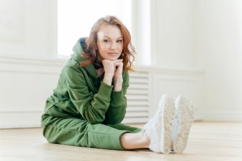 Young slim young woman keeps hands under chin, has make up, sits on floor and stretches legs, wears green sweatsuit, looks straightly at camera. People, workout and pilates exercises concept