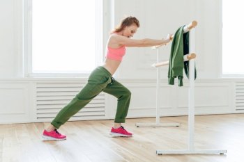 Sport, recreation, flexibility concept. Motivated strong female makes plank, leans at ballet barre, stretches before exercises prepares muscles or biceps for hard training. Aerobics instructor indoor