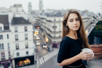 Pretty dreamy woman with mug standing on balcony and looking away in Paris, France. . Dreamy woman with mug on balcony