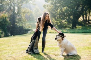 Beautiful young woman with parka standing on grass and touching hat on head of her dog.. Woman touching hat on head of dog