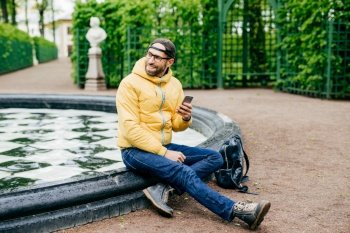 Relaxed carefree bearded traveler male in trendy cap, anorak, jeans and eyewear having excursion in forest reserve sitting near fountain holding smartphone typing messages looking aside with smile