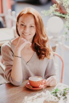 People and free time concept. Positive red haired woman with gentle smile, keeps hand under chin, dressed in casual jumper, drinks aromatic coffee in outdoor cafeteria, enjoys talk with friend