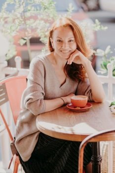 Vertical shot of cheerful foxy female dressed in fashionable clothes, drinks coffee, poses in outdoor cafe, enjoys spare time, has pleasant smile on face, keeps hand under chin. Lifestyle concept