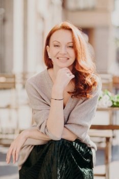 Cheerful attractive red haired young woman has positive expression, keeps hand under chin, wears fashionable attire, looks direclty at camera, poses outside, being in high spirit, smiles happily