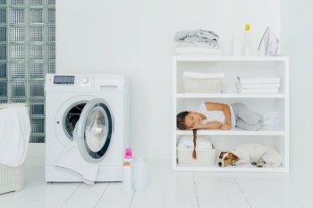 Tired female child rests on console shelf together with pet, does washing at home, sleeps in laundry room, opened washing machine with dirty towel inside. Childhood, cleanliness, family chores concept