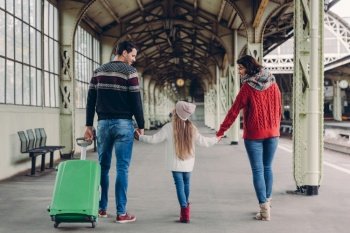 Horizontal view of affectionate parents and their dauhter keep hands together, going to have trip abroad, pose on railway station, carry green bag, have happy looks, communicate. Family journey