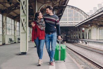 Young family couple embrace and look at each other with smile, wear jeans and warm sweaters, carry suitcase, pose on railways station meet after long departure. Boyfriend and girlfriend cuddle outside
