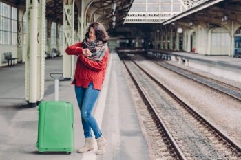 Photo of attractive young woman waits train at railway station, dressed in knitted sweater and jeans, stands near green bag, looks at watch. Pleased passenger with lugagge has business trip.