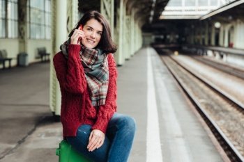 Beautiful brunette female wears knitted sweater and scarf, sits on suitcase near platform, communicates on smart phone, feels bored to wait for long time. People, travelling and waiting concept
