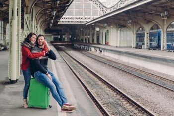 People, travelling, relationship concept. Happy unshaven husband and wife embrace at railway station, going to have trip, have vacation. Handsome man speaks on smart phone, recieves hug girlfriend