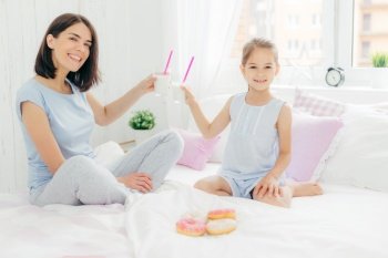 Horizontal shot of pretty female and her daughter clink glasses with cocktail in bed, have breakfast in bedroom, eat delicious doughnuts, look happily at camera. Young mother and her kid at home