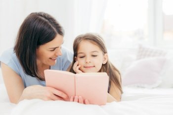 Affectionate young mother with dark hair, looks happily at her small daughter, read interesting book or fairy tale together, enjoy domestic atmopshere at home. People, family, childhood concept