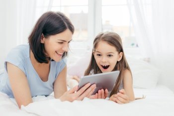 Beautiful mother and daughter watch something funny on tablet computer, connected to wireless internet, spend free time in bedroom, have surprised happy expression. People and leisure concept