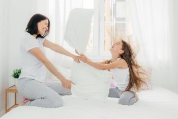 Parenthood and family concept. Attractive female mother with positive smile, spends free time with her daughter, have pillow fight, pose against bedroom interior, enjoys leisure time during weekend