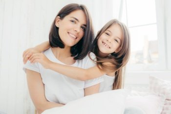 Portrait of happy Caucasian mother with charming smile and her small daughter embraces with love mum, being in good mood, pose on comfortable bed against cozy interior. People and family concept