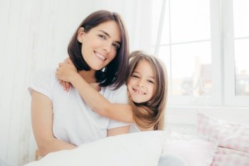 Positive daughter and mum embrace each other, being in high spirit after good sleep, pose in bedroom, smile gently on face, have pleasant appearance. Pretty child with her mother. Morning and bed time