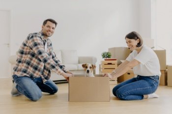 Positive millennial couple play with favourite pet, have fun during moving in new apartment, stand on knees near carton box with dog, enjoy domestic atmosphere, live together in modern house