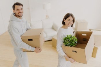 Positive female and male property owners pose with personal belongings in carton boxes, relocate to own apartment, smile pleasantly, pose in white living room. Moving day, planning, relocation