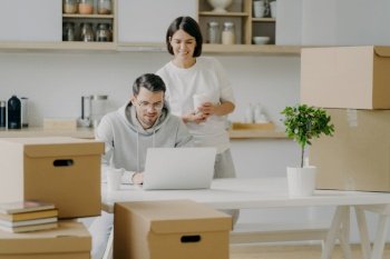 Photo of cheerful young woman stands behind her husband who works on laptop computer, pose in modern kitchen of their new apartment, surrounded with cardboard boxes, think about modern design