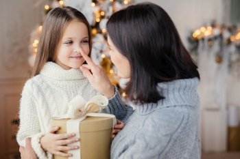 Affectionate mother gives present to her adorble little daughter, prepares surprise on Christmas, touch her nose, expresses great love. Family, celebration, presents, miracle, winter holidays concept