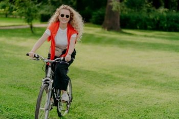 People, nature, rest, lifestyle concept. Happy curly woman rides bicycle among green grass, moves actively, wants to be fit, explores new places in countryside, wears sunglasses, casual clothes