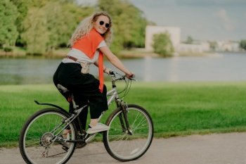 Image of female model rides bicycle, looks aside with cheerful expression, wears sunglasses, breathes fresh air, poses near lake and green trees, covers long distance, finds out something new