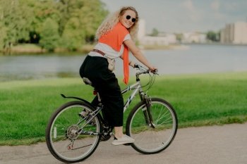Pleased young woman enjoys new route on bicycle, rides among lake, green lawn and buildings far away into distance, wears summer shades, casual outfit, white sneakers, being in good physical shape