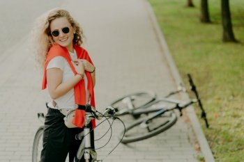 Cheerful curly haired female stops during way, poses on bicycle at road, friends bike lies near, wears sunglasses, t shirt and trousers, carries little bag around waist, being fit and healthy