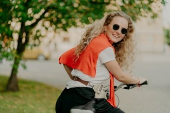 Photo of delighted woman poses back at camera, rides bicycle outdoor, wears sunglasses, t shirt and sweater on shoulders, poses near green tree, has toothy smile positive look carries bag with herself