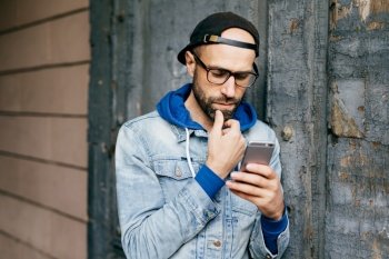 Serious concentrated bearded man in cap and denim jacket standing against cracked wall holding smartphone liking posts and sharing them via social networks, reading news with deep and thoughtful look