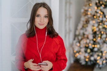 Horizontal shot of young Caucasian woman enjoys music from playlist, listens favourite song with earphones, dressed in knitted red sweater, stands against decorated Christmas tree background