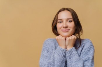 Lovely dark haired woman with healthy skin, keeps both hands under chin, wears oversized sweater, looks directly at camera, isolated over brown studio wall. People, human facial expressions.