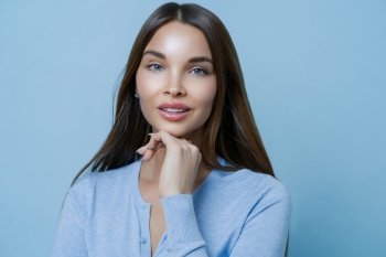 Beauty is in natural look. Portrait of pleasant looking European woman keeps hand under chin, looks with calm expression at camera, consults beautician, isolated over blue studio background.