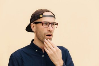 Sideways shot of handsome man with stubble looks with puzzlement aside, keeps hand on chin, wears spectacles and stylish black cap, isolated over beige background. People and emotions concept