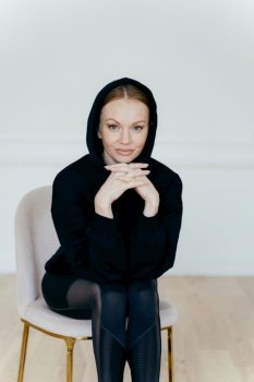 Satisfied confident woman with minimal makeup, wears black hoody, keeps hands under chin, looks at camera, sits on comfortable chair, poses indoor. Sporty female coach has rest. Vertical shot