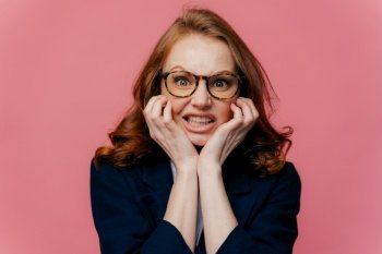 Negative feeling concept. Dissatisfied redhead prosperous businesswoman clenches teeth from irritation, keeps hands under chin, wears transparent glasses and black formal outfit, isolated on pink wall
