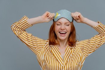 Overjoyed red haired Caucasian woman laughs positively, wears blindfold and yellow striped pajama, expresses good emotions, isolated on grey background. Good rest, bedtime and sleeping concept