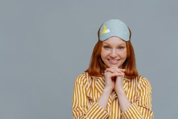 Pretty lady with ginger hair, keeps hands under chin, looks directly at camera, wears blindfold on head, striped yellow pyjama, has good rest, isolated over grey background, copy space for your text