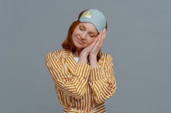 Pleased cheerful woman wears blindfold and striped pajamas, has happy expression, awakes in good mood, has healthy sleep habits, feels totally relaxed, smiles broadly, isolated on grey background