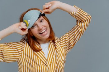 Pleased cheerful woman wears blindfold and striped pajamas, has happy expression, awakes in good mood, has healthy sleep habits, feels totally relaxed, smiles broadly, isolated on grey background