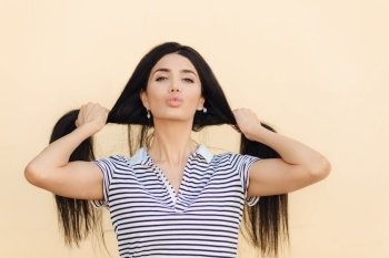 Lovely young Caucasian female shows her long straight dark hair, pouts lips, demonstrates natural beauty, poses against light background, dressed in striped t shirt. Hair care and beauty concept