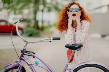 Horizontal shot of happy woman rides bicycle in park, has break, poses at camera, keeps mouth opened, touches cheeks, wears trendy sunglasses, has curly red hair, enjoys nice weekend during summer