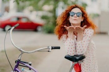 Affectionate redhead woman sends air kiss to lover on street, keeps palms near mouth, wears round shades, dress, leans at bicycle, has active rest outdoor, poses on street. Recreational time concept