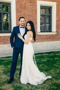 Vertical portrait of attractive bride and handsome bridegroom stand close to each other, hug passionately. Cheerful young wedding couple stand outdoor on green grass, have happy delightful look