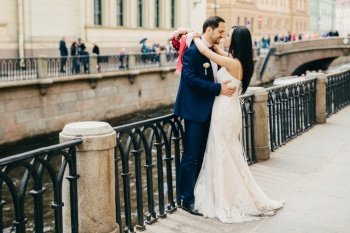Brunette female in white dress embraces her bridegroom, looks with great love at his eyes, stand together at bridge. Future family celebrate first day of their marriage, have good relationships