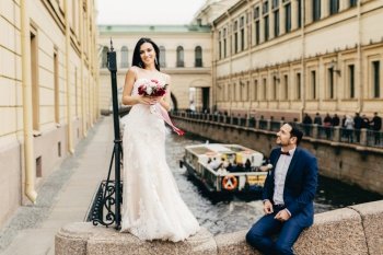 Cute adorable bride with long dark hair, wears wedding dress, holds beautiful bouquete of flowers, stands on bridge and her husband who sits near her, looks with love and passion. Romance concept