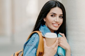 Outdoor shot of brunette female student with makeup, dark long hair, dressed in denim jacket, holds takeaway coffee, textbook, carries rucksack, looks somewhere into distance, strolls in city